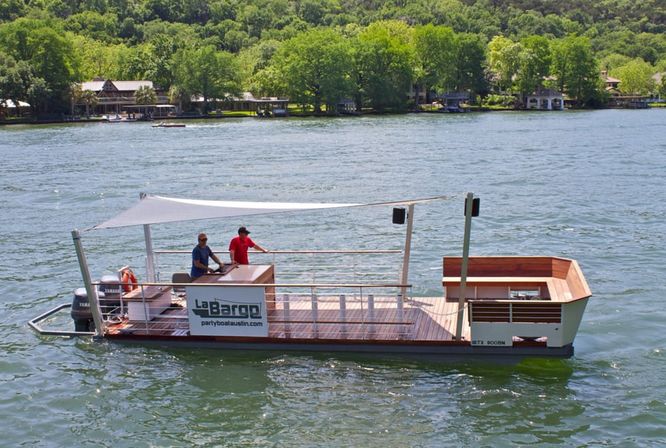 Sunlit wooden party pontoon boat with white canopy and two people aboard cruising on a tree-lined lake in front of waterfront homes.