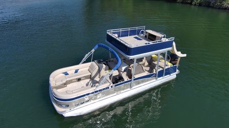 Aerial view of a blue-and-white double-decker pontoon boat with upper deck and slide floating on calm green lake water near a tree-lined shore