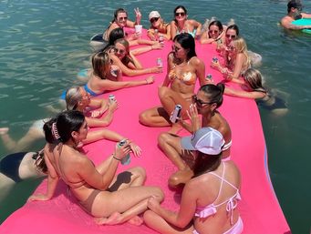 Sun-soaked group of women in bikinis lounging and sipping canned drinks on a large pink floating mat in a lake, chatting and relaxing on a summer day.