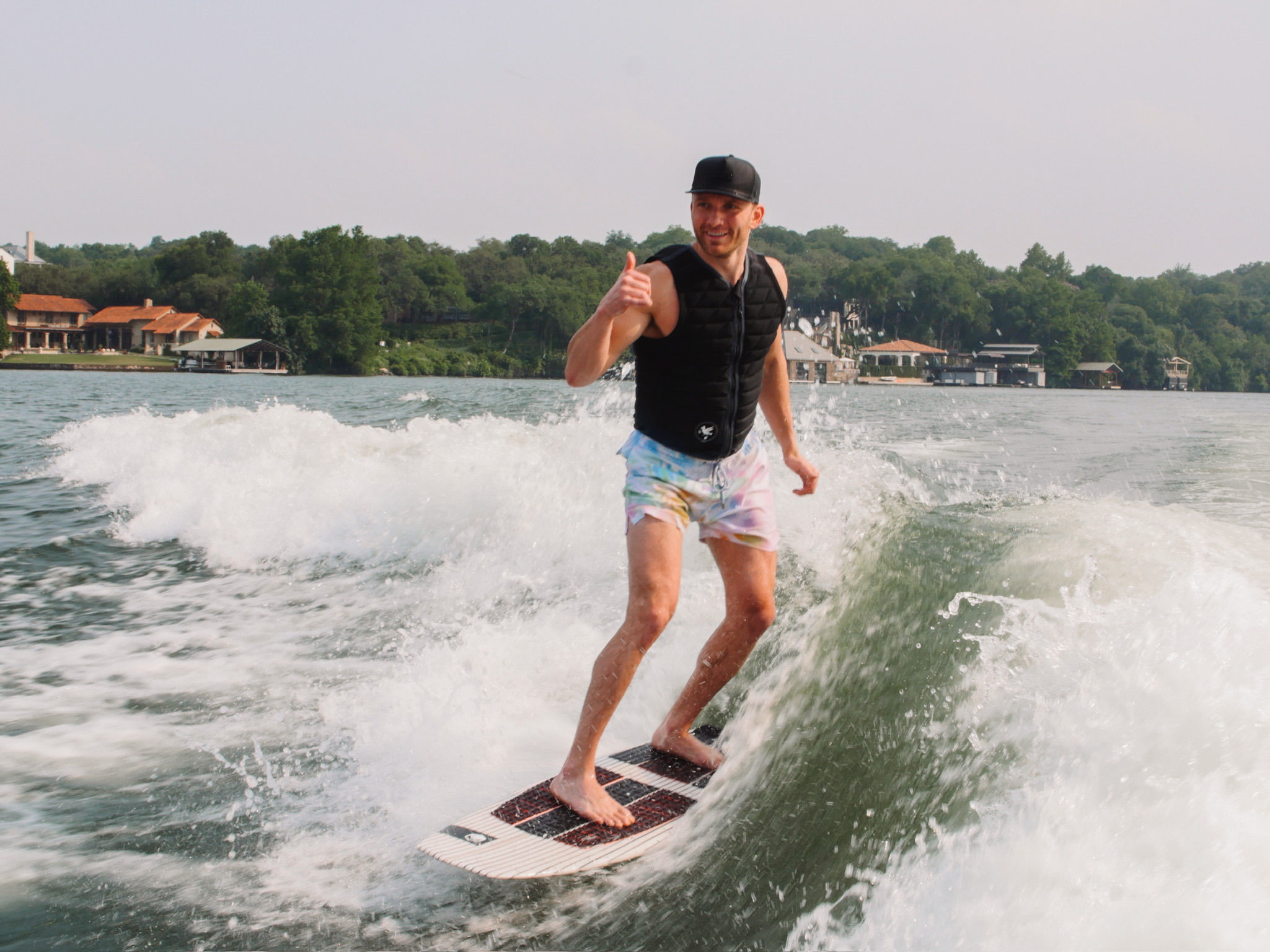 Man wakesurfing on a lake near a tree-lined shoreline and lakeside homes, wearing a black life vest, cap and pastel shorts while giving a thumbs-up