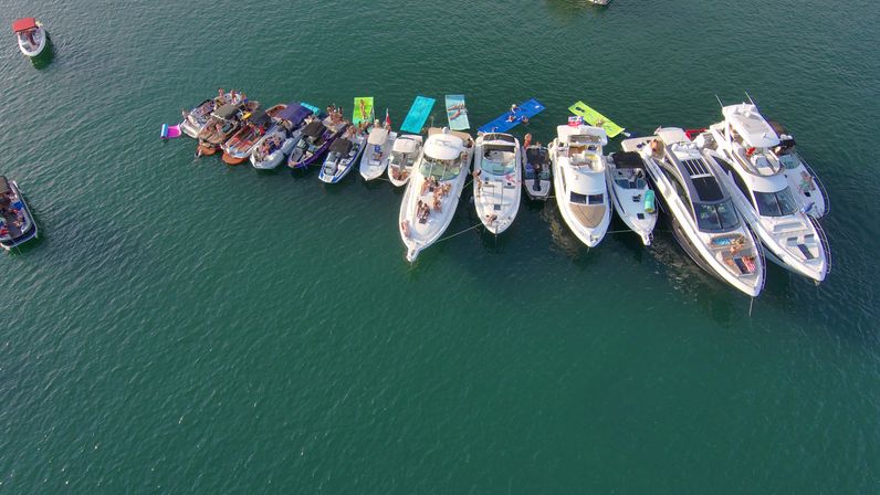 Aerial view of yachts and small boats anchored side-by-side in a sunny coastal bay with colorful floating mats and people relaxing onboard
