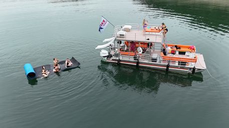 Pontoon party boat with orange cushions anchored on a calm lake; people lounging on upper and lower decks with a slide, while four friends float nearby on a black mat and blue foam roller.