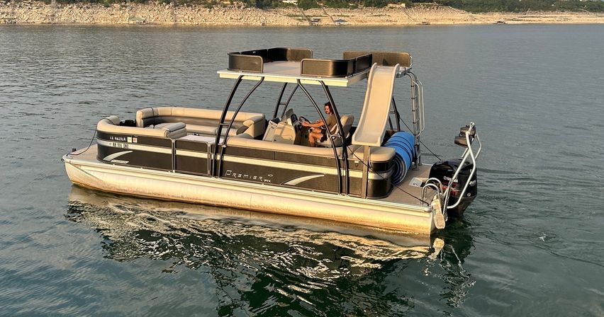 Pontoon boat with rooftop lounge and water slide, person steering on a calm lake at golden hour near a rocky shoreline — summer-ready boating scene.