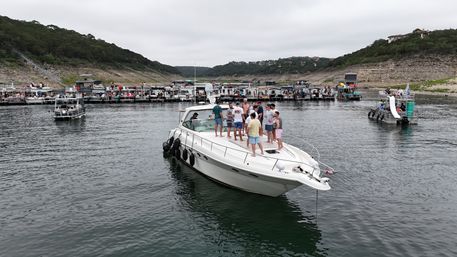 Group of people socializing on a white motor yacht near a crowded lake marina with pontoon boats, rocky tree-covered shoreline, and overcast sky