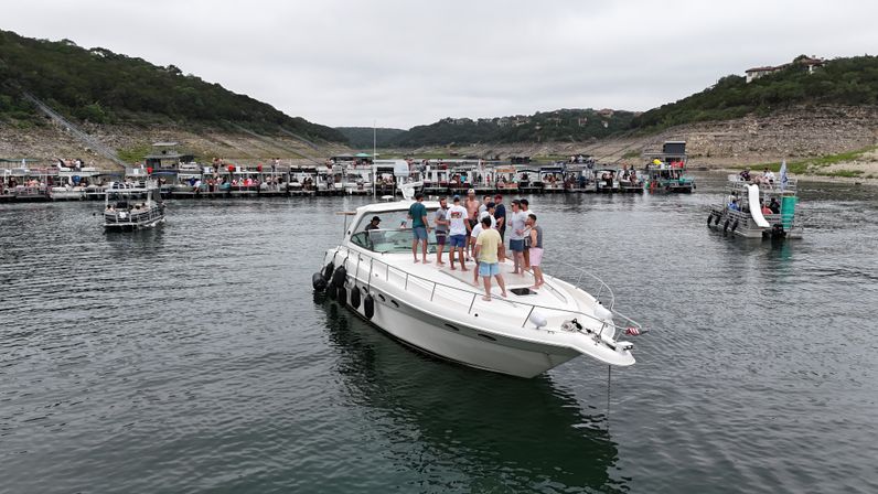 Group of people socializing on a white motor yacht near a crowded lake marina with pontoon boats, rocky tree-covered shoreline, and overcast sky