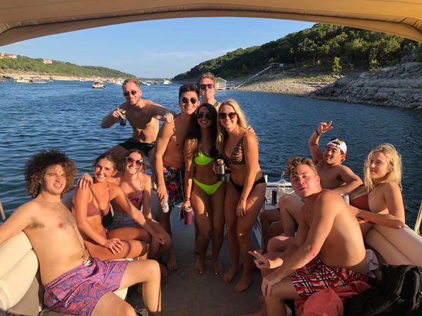 Sunlit summer lake party on a pontoon boat – a group of friends in swimsuits smiling and holding drinks with wooded hills and a rocky shoreline in the background.