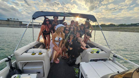 Sunset lake party on a pontoon boat: a smiling group of women in swimsuits and sun hats posing under a canopy with drinks and coolers, calm water and a cloudy shoreline in the background.