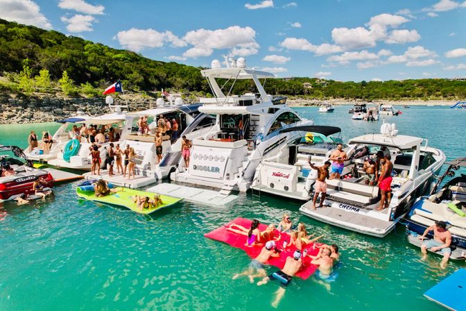 Aerial view of a lively summer raft-up: multiple yachts anchored side-by-side on turquoise lake water, people socializing on swim platforms and colorful floats, wooded shoreline and sunny blue sky.