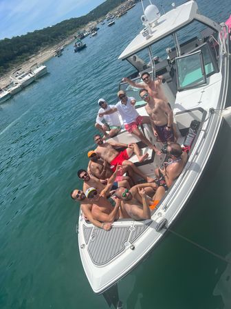 Group of friends enjoying a boat party on a white motorboat in clear turquoise water on a sunny day, anchored near a rocky shoreline with other boats in the background.
