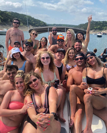 Cheery summer boat party: a large group of adults in swimwear smiling and holding drinks on a crowded boat on a sunny lake with tree-covered hills