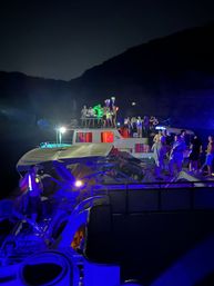 Nighttime lake boat party with multiple anchored boats and pontoons, colorful LED lights and glow sticks, people dancing beneath dark mountain silhouettes.
