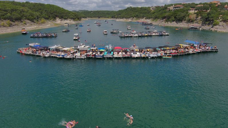 Aerial view of a lively lake party: dozens of linked pontoon boats forming a crowded floating raft on green-blue water, swimmers nearby and tree-covered shoreline in the background.