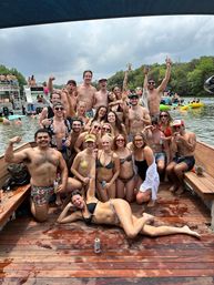 Large group of friends in swimsuits posing and cheering on a wooden lake dock at a lively summer boat party with inflatables and boats