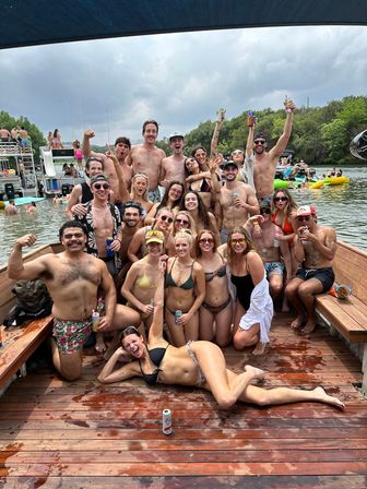 Large group of friends in swimsuits posing and cheering on a wooden lake dock at a lively summer boat party with inflatables and boats