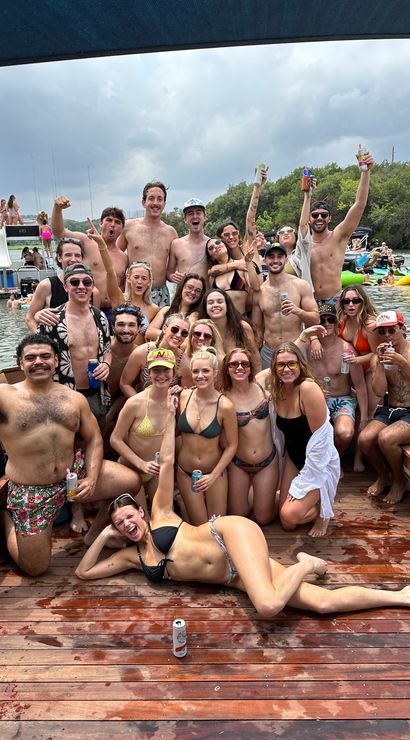 Large group of friends in swimsuits posing and cheering on a wooden lake dock at a lively summer boat party with inflatables and boats