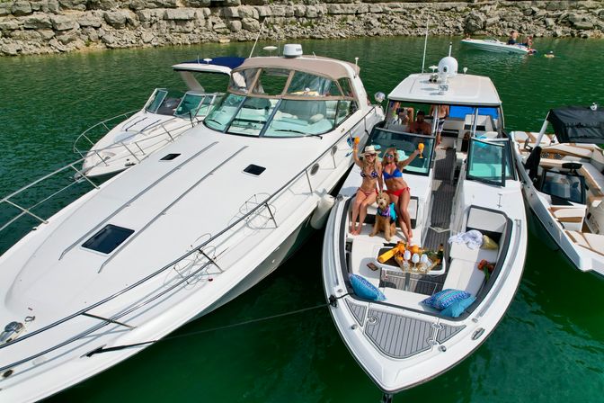 Two women in bikinis with a dog raising drinks on the bow of a moored motorboat between yachts for a summer lake boat party near a rocky shoreline, tray of champagne and glasses visible