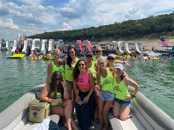 Group of women in neon tank tops smiling on a pontoon boat at a busy summer lake party with inflatable slides, colorful floats, many boats, an American flag and wooded shoreline under a partly cloudy sky