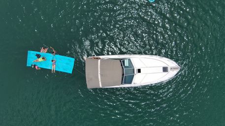 Drone view of a white motorboat anchored in green water with people lounging on a bright blue floating mat tied to the stern — summer boating fun.