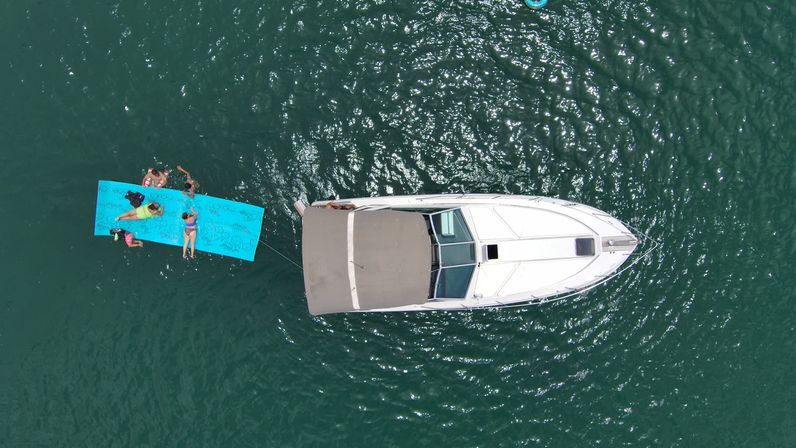 Drone view of a white motorboat anchored in green water with people lounging on a bright blue floating mat tied to the stern — summer boating fun.