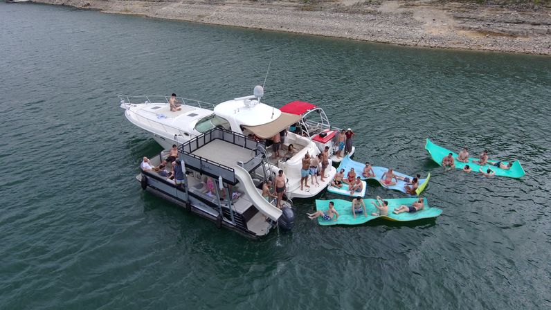 Lake boat party: a yacht and attached pontoon with a slide anchored near a rocky shoreline while a group lounges and swims on bright teal floating mats in green water.