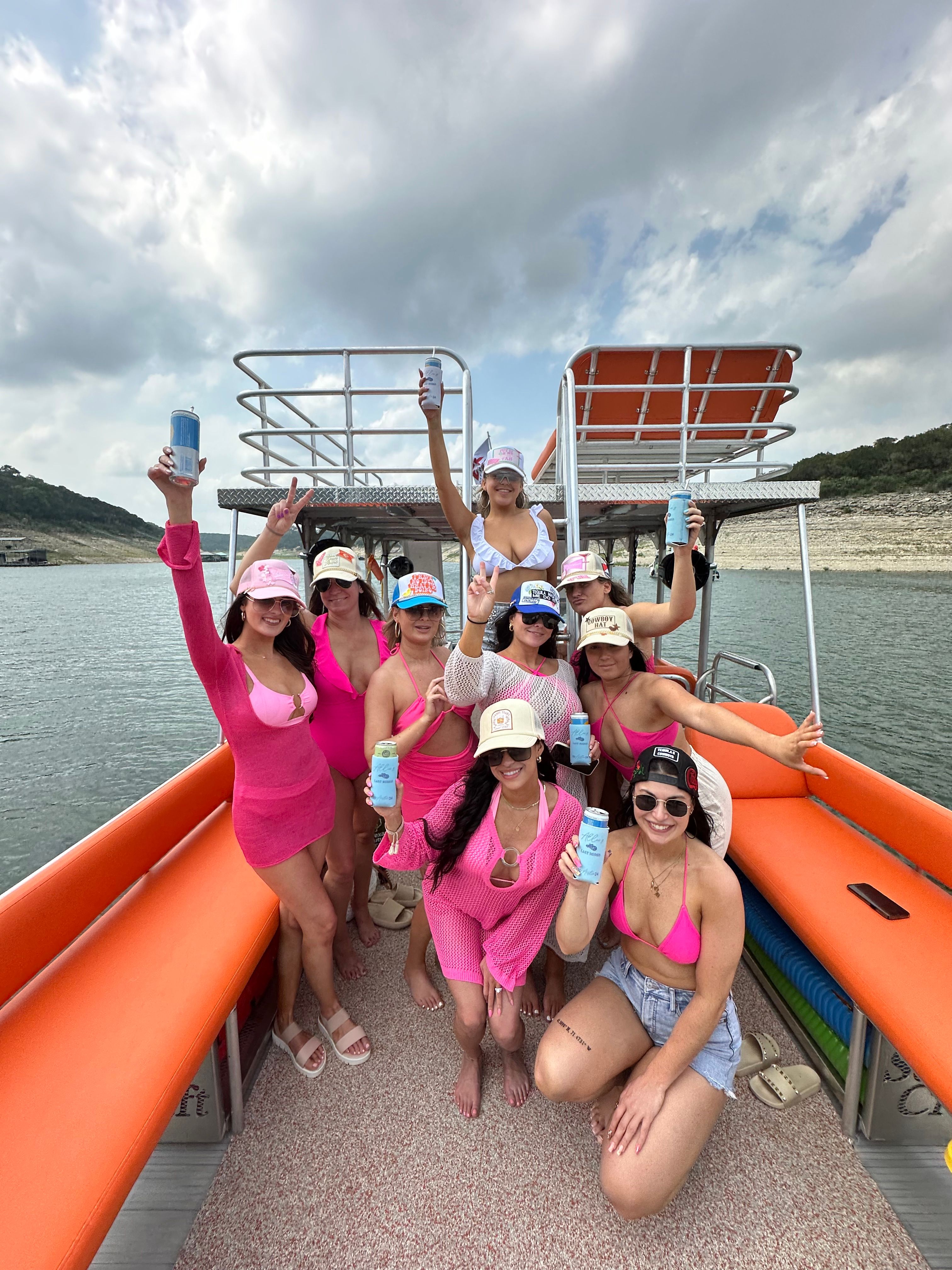 Group of women in pink swimsuits and hats cheering with canned drinks on an orange-seated pontoon boat — lake day with rocky shoreline and cloudy summer sky.