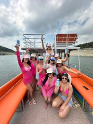Group of women in pink swimsuits and hats cheering with canned drinks on an orange-seated pontoon boat — lake day with rocky shoreline and cloudy summer sky.