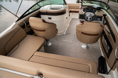Cozy tan-upholstered motorboat interior with two swivel captain chairs facing a helm with steering wheel and gauges, L-shaped bench seating and textured deck, docked at a calm marina.