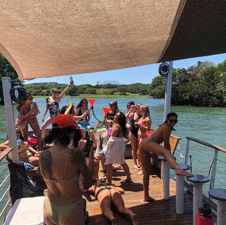 Summer lake party on a shaded pontoon: a group of friends in swimsuits drinking from colorful funnels, dancing, sunbathing and laughing with a green shoreline and calm water in the background.