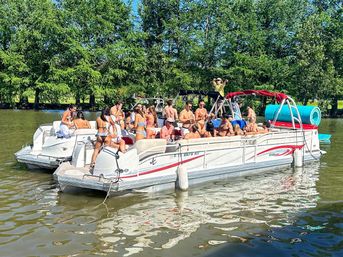 Sunny pontoon boat party on a calm, tree-lined river with a group of people in swimsuits, a blue inflatable tube and relaxed summer vibes.