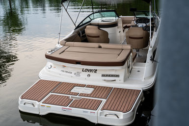Rear view of a white sundeck motorboat docked on calm water, with a tan cushioned sun pad, faux-teak swim platform, twin helm seats and an open cockpit ready for cruising.