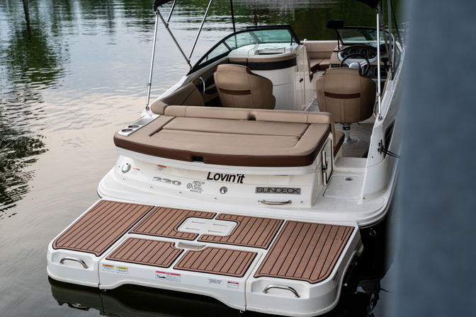 Rear view of a white sundeck motorboat docked on calm water, with a tan cushioned sun pad, faux-teak swim platform, twin helm seats and an open cockpit ready for cruising.