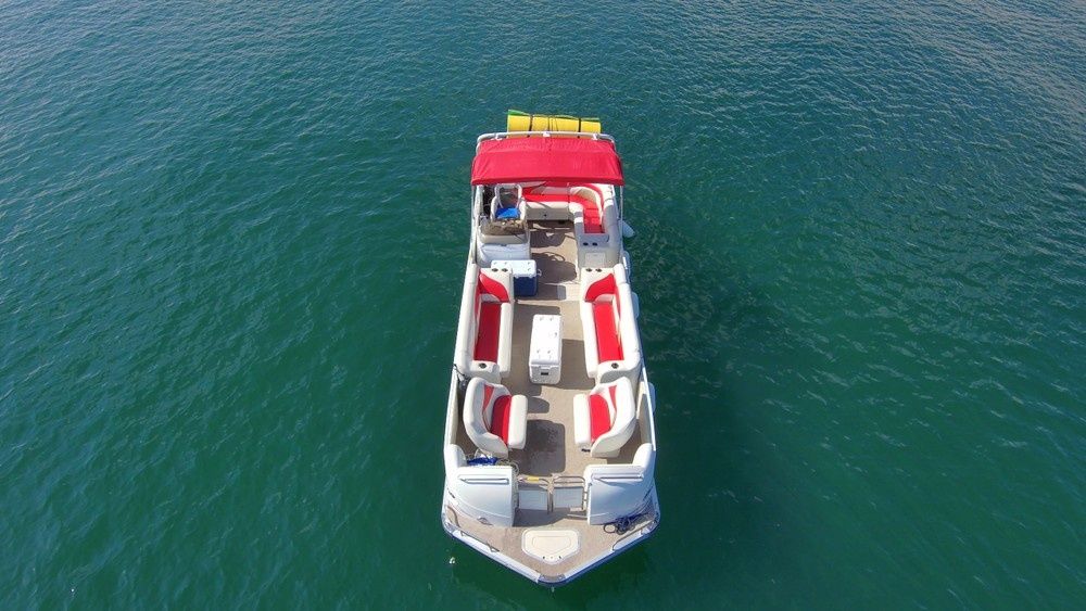 Aerial view of a red-and-white pontoon boat with canopy and cushioned seating floating on calm teal lake water, ready for a summer cruise.
