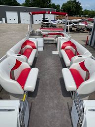 Pontoon boat interior with red-and-white cushioned bench seating, walk-through center aisle on gray carpeted deck, red bimini top and cupholders, parked at a marina with stored boats and storage building in background