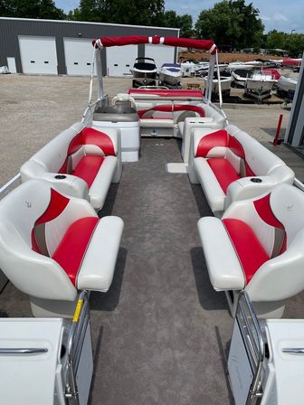 Pontoon boat interior with red-and-white cushioned bench seating, walk-through center aisle on gray carpeted deck, red bimini top and cupholders, parked at a marina with stored boats and storage building in background