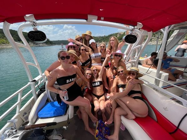 Group of women in matching 'BABE' swimsuits and one 'BRIDE' top smiling and posing on a red-canopied pontoon boat on a sunny lake, wearing sunhats and sunglasses and holding drinks — summer bachelorette boat party.