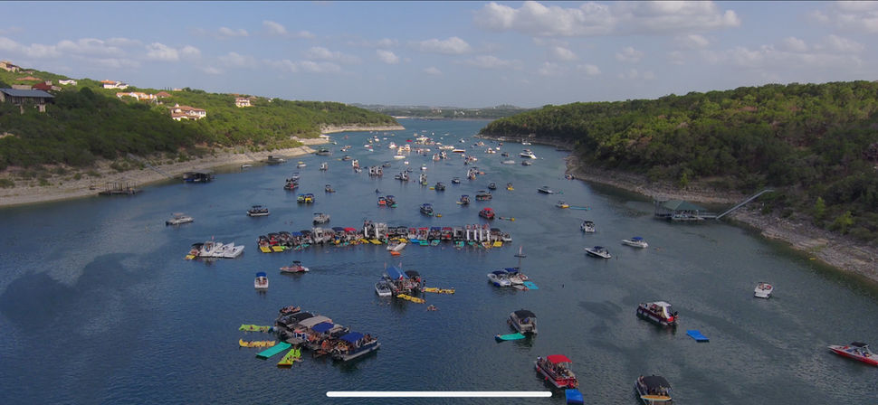 Aerial view of a busy lake cove with dozens of boats, pontoons and colorful floating platforms clustered in a calm blue reservoir, framed by wooded hills and a partly cloudy sky.