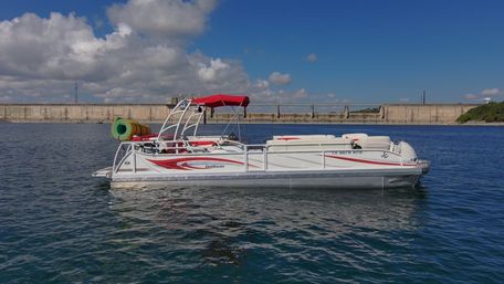 White-and-red pontoon boat with red canopy and colorful pool floats drifting on calm blue reservoir water near a concrete dam under a sunny, partly cloudy sky.