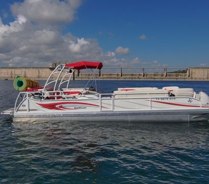 White-and-red pontoon boat with red canopy and colorful pool floats drifting on calm blue reservoir water near a concrete dam under a sunny, partly cloudy sky.