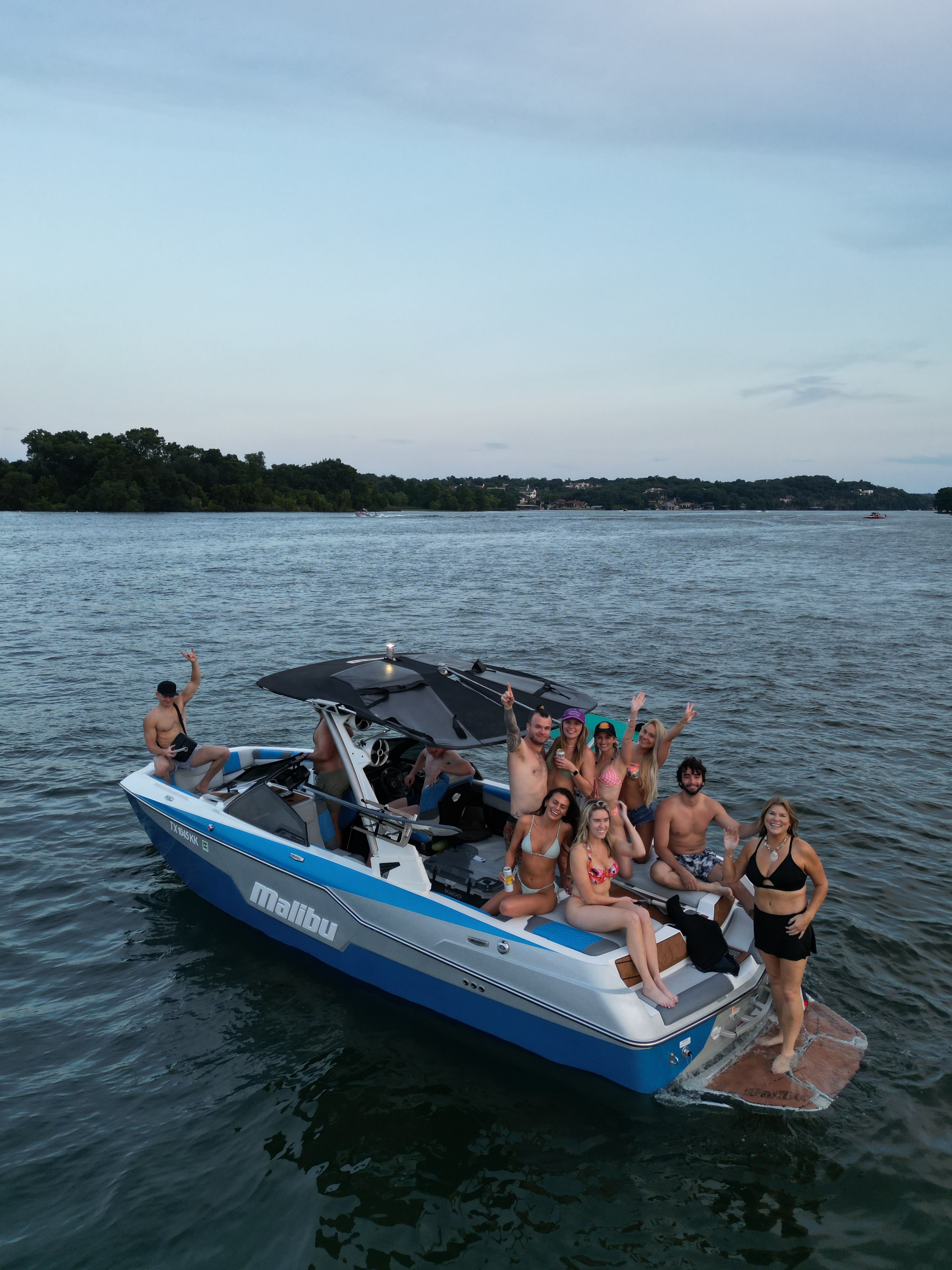 Group of friends in swimsuits waving and holding drinks on a blue boat anchored on a calm lake at dusk with a tree-lined shore in the distance.