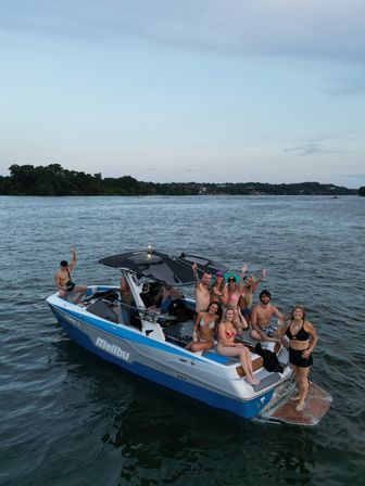 Group of friends in swimsuits waving and holding drinks on a blue boat anchored on a calm lake at dusk with a tree-lined shore in the distance.