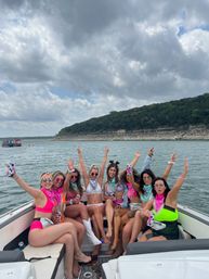 Smiling group of friends in colorful swimsuits and bandanas raising hands and drinks while partying on a motorboat on a lake with a wooded rocky shoreline and dramatic cloudy sky