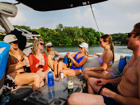 Group of friends in swimsuits on a speedboat at a sunny lake, popping champagne and sipping drinks with a wooded shoreline in the background.