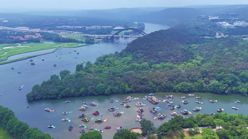 Aerial lake view of a crowded cove with colorful party boats and rafts, forested shoreline, and an arched road bridge spanning the river in the distance.