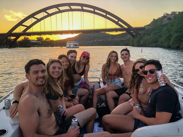 Sunset boat party with a group of friends in swimsuits holding canned drinks on a lake, smiling together beneath a large arched bridge and wooded shoreline.
