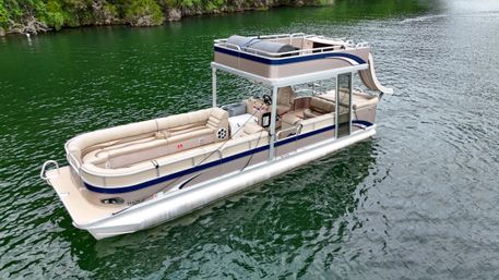 Beige double-deck pontoon boat with upper deck and slide floating on a green lake near a tree-lined shore