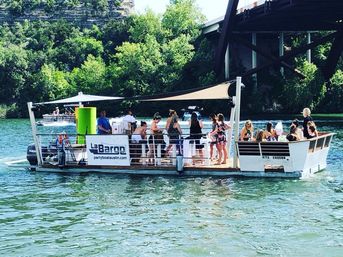 Sunny party pontoon on Lake Austin with a group of people under a canopy, passing a bridge and green limestone bluffs.