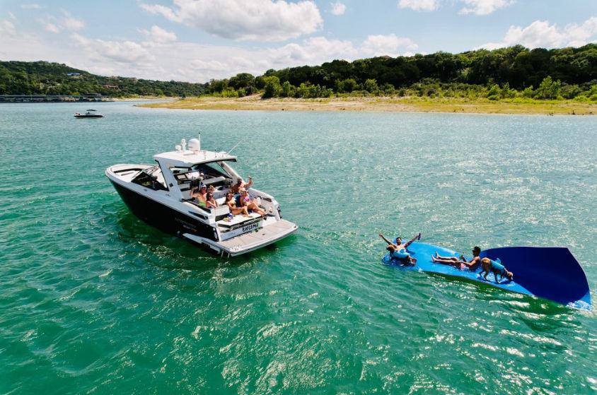 Friends on a recreational motorboat enjoying a sunny day on turquoise lake water, lounging on the stern and playing on a large blue inflatable water mat near a tree-lined shoreline