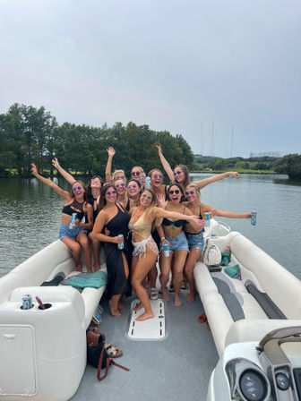 Cheerful group of women in swimsuits and sunglasses partying on a pontoon boat on a calm lake, holding canned drinks, laughing and waving with tree-lined shore and cloudy sky behind them.