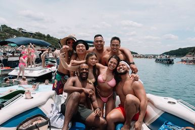 Group of friends in swimsuits smiling and posing on a motorboat at a sunny summer lake boat party, with many other boats and people on the water in the background.