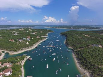 Aerial drone view of a summer lake cove with dozens of boats clustered on turquoise water, surrounded by tree‑covered shores and waterfront homes beneath a bright blue sky